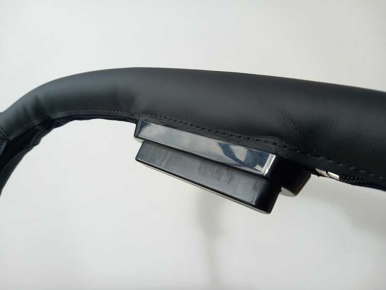 May include: Close-up of a black leather steering wheel cover with a black plastic control panel attached.