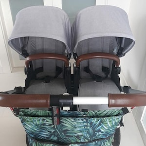 May include: A double stroller with gray fabric seats and brown leather handles. The stroller has a large, tropical-print bag attached to the bottom.