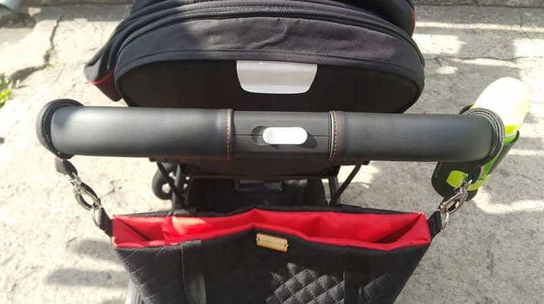 Puede incluir: Cochecito negro con una bolsa roja y negra adjunta. El cochecito tiene un asa de cuero negro con un bot&oacute;n blanco. Hay un portavasos verde y negro adjunto al asa.