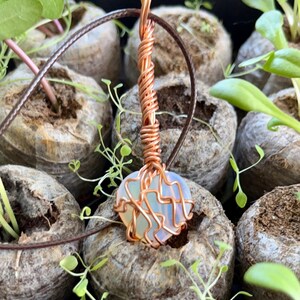 May include: A copper wire wrapped heart pendant with a white opal stone. The pendant is hanging from a brown leather cord.