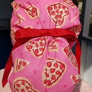 May include: A pink scrub cap featuring a repeating pattern of pizza slices with heart-shaped pepperoni. The cap is tied with a bright red ribbon. The fabric has a subtle glitter effect.