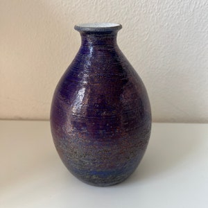 Puede incluir: Un pequeño jarrón de cerámica de color morado oscuro con un borde blanco. El jarrón tiene una superficie texturizada, ligeramente rugosa.