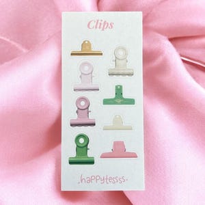 Op de afbeelding: Een kaart met het woord "Clips" en de tekst "-happytessss-" toont acht kleurrijke bindclips in goud, roze, groen en crème. De clips zijn gerangschikt in twee kolommen van vier op een witte achtergrond, tegen een roze satijnen stof.