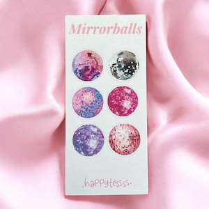 Può includere: Un foglio di adesivi rettangolare con sei disegni di sfere a specchio colorate. Il testo "Mirrorballs" è stampato in alto e ".happyteessss." in basso. Lo sfondo è un tessuto di raso rosa.