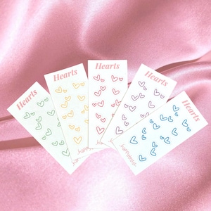 Op de afbeelding: Vijf witte kaarten met het woord "Hearts" en kleine hartillustraties in verschillende kleuren. De kaarten zijn gerangschikt op een roze satijnen oppervlak. De tekst ".happytessss." is zichtbaar.