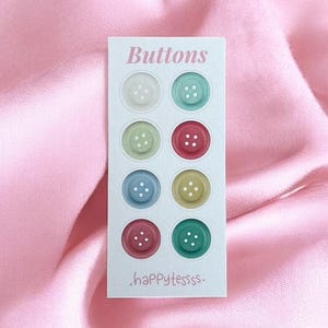 Op de afbeelding: Een kaart met acht kleurrijke knopen in verschillende tinten wit, groen, rood, blauw en geel. De kaart is gelabeld met "Buttons" en heeft de tekst ".happytessss." onderaan. De knopen zijn geplaatst tegen een roze satijnen achtergrond.