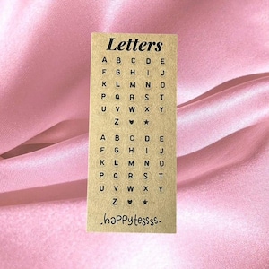 Op de afbeelding: Een rechthoekige kaart van kraftpapier met bovenaan het woord "Letters". De kaart bevat het alfabet, waarbij elke letter in een klein, zwart lettertype is gedrukt. De onderkant van de kaart luidt ".happytessss." in een handschriftstijl. De achtergrond is een roze satijnen stof.