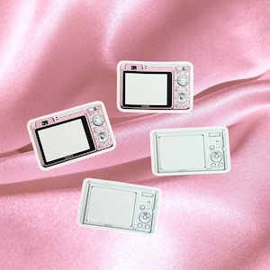 Puede incluir: Cuatro pequeñas cámaras digitales rosas y blancas están dispuestas sobre un fondo de satén rosa. Dos cámaras tienen pantallas negras, mientras que las otras dos tienen pantallas blancas. Las cámaras tienen botones y diales.