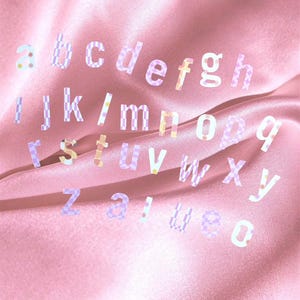 May include: A collection of lowercase alphabet letters in a variety of pastel colors and patterns. The letters are scattered across a pink satin fabric, creating a soft and dreamy aesthetic. The letters are in a variety of fonts.