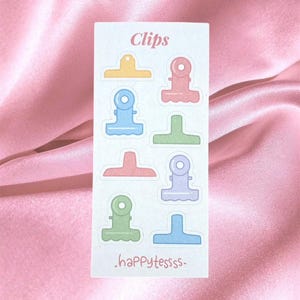 Op de afbeelding: Een rechthoekig stickervel met pastelkleurige illustraties van klemmen in geel, roze, blauw, groen en paars. De tekst "Clips" staat bovenaan en de tekst "happytessss" staat onderaan op het witte vel gedrukt.