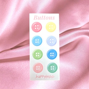 Op de afbeelding: Een rechthoekige witte kaart met acht ronde knopen in pasteltinten zoals roze, geel, blauw, lavendel, lichtgroen en taling. Het woord Buttons staat bovenaan in een schreeflettertype en de tekst happytessss staat onderaan de kaart.