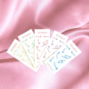 Op de afbeelding: Vijf rechthoekige stickers met het woord "Arrows" bovenaan gedrukt. Elke sticker heeft een ander gekleurd pijldesign, waaronder groen, rood, paars, geel en blauw. De stickers zijn gerangschikt op een roze satijnen oppervlak.