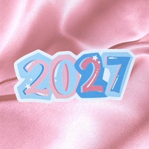 Op de afbeelding: Een gestanste sticker met het jaartal 2027 in een gestileerd lettertype met bubbelletters. De cijfers zijn gekleurd in tinten lichtroze en blauw met witte accenten en kleine sterretjes, wat zorgt voor een gelaagd, driedimensionaal uiterlijk.