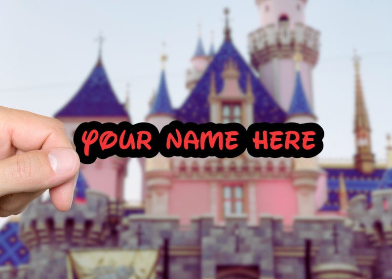K&ouml;nnte beinhalten: Ein schwarzer Aufkleber mit dem Text "YOUR NAME HERE" in roter, stilisierter Schrift. Der Aufkleber wird vor einem verschwommenen Hintergrund mit einem rosa Schloss mit blauen T&uuml;rmen gehalten, was eine Themenpark-Atmosph&auml;re hervorruft.
