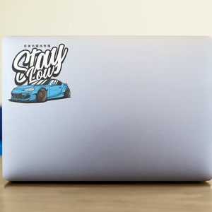 Peut inclure: Un ordinateur portable blanc avec un autocollant représentant une voiture bleue avec l'inscription "Stay Low" en lettres blanches.