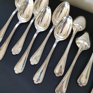 Puede incluir: Un conjunto de cucharas plateadas dispuestas sobre una superficie oscura. Las cucharas tienen un acabado pulido y reflectante, con cuencos alargados y bordes decorativos festoneados en los mangos. Probablemente son parte de un juego de comedor.