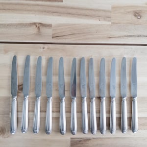 Op de afbeelding: Een rij van twaalf zilveren messen op een lichtbruin houten oppervlak. Elk mes heeft een lang, slank lemmet en een decoratief handvat met een afgeronde basis. De messen zijn gelijkmatig verdeeld en reflecteren het licht.
