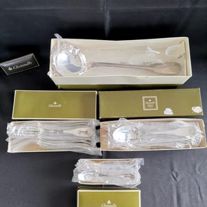 Puede incluir: Juego de cubiertos Christofle plateados, que incluye un cucharón, cucharas y tenedores, empaquetados en cajas verdes y blancas. El logotipo de Christofle es visible en las cajas y en una tarjeta separada. Los cubiertos se muestran sobre una superficie negra.