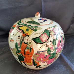 Puede incluir: Un tarro de cerámica redondo, color crema, con tapa, que presenta una escena pintada a mano de figuras, dragones y elementos florales en rojo, verde, rosa y amarillo. La tapa tiene un pequeño pomo rojo y blanco.