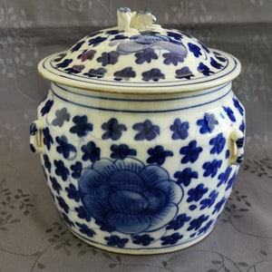 Puede incluir: Un tarro de porcelana blanca con un diseño floral azul y blanco. El tarro tiene una tapa con un pequeño pomo en la parte superior. El tarro está decorado con una gran flor azul y flores azules más pequeñas.