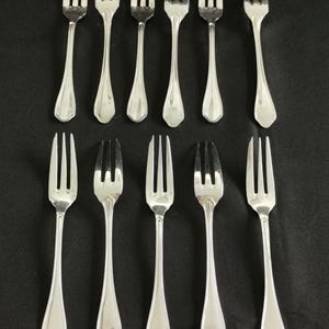 Pode incluir: Uma coleção de doze garfos prateados dispostos sobre uma superfície preta. Cada garfo tem três dentes e um cabo longo e fino com uma forma decorativa. Os garfos estão uniformemente espaçados, mostrando o seu design elegante.