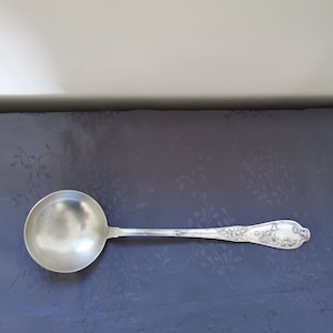 Puede incluir: Un cucharón de plata con un cuenco redondo y un mango largo y ornamentado. El mango presenta diseños detallados de flores y volutas. El cucharón probablemente está hecho de plata de ley y está diseñado para servir sopas o salsas.