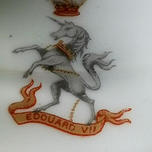 Puede incluir: Superficie blanca con borde dorado e ilustración detallada. La ilustración presenta un unicornio, una corona y una pancarta que dice "EDOUARD VII" en letras rojas. El unicornio es gris con un collar dorado.