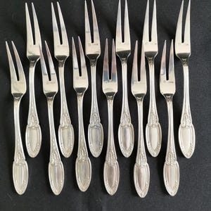 May include: A collection of silver-colored forks arranged on a black surface. Each fork has a three-tine design and an ornate handle. The forks are evenly spaced, showcasing their elegant design.
