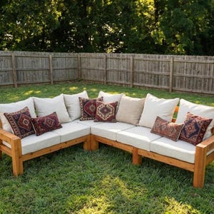 May include: An L-shaped outdoor sectional sofa constructed from natural wood with thick, cream-coloured seat cushions. The sofa is decorated with several large cream-coloured back cushions and smaller rectangular accent pillows featuring dark red patterns.