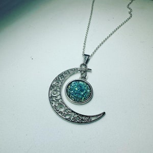 K&ouml;nnte beinhalten: Eine silberne Halskette mit einem Halbmond-Anh&auml;nger. Der Mond ist aus filigranem Metall gefertigt und hat einen runden, mit blauen Glitzer gef&uuml;llten Cabochon in der Mitte.