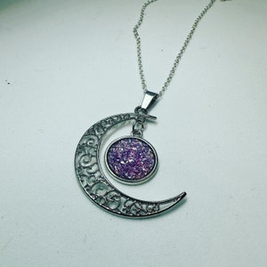 K&ouml;nnte beinhalten: Eine silberne Halbmonds-Halskette mit einem filigranen Design. Der Anh&auml;nger hat einen runden, mit violettem Glitzer gef&uuml;llten Cabochon.