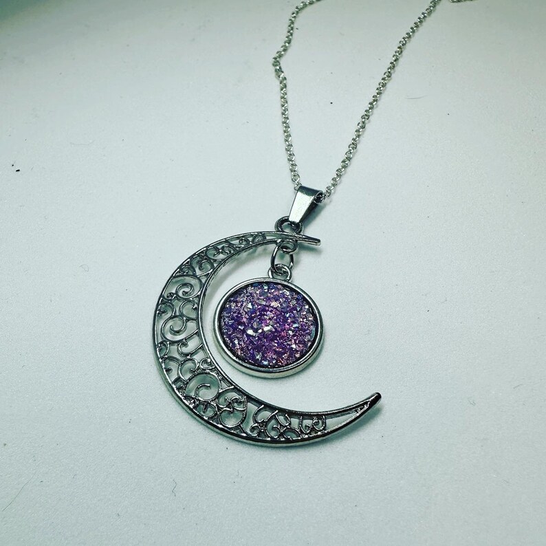 K&ouml;nnte beinhalten: Eine silberne Halbmonds-Halskette mit einem filigranen Design. Der Anh&auml;nger hat einen runden, mit violettem Glitzer gef&uuml;llten Cabochon.