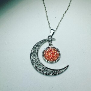 K&ouml;nnte beinhalten: Eine silberne Halskette mit einem Halbmond-Anh&auml;nger mit filigranem Design. Der Halbmond-Anh&auml;nger hat eine runde, rote, glitzernde Mitte.