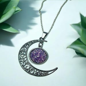 K&ouml;nnte beinhalten: Eine silberne Halbmonds-Halskette mit einem Anh&auml;nger, der mit violettem Glitzer gef&uuml;llt ist. Der Anh&auml;nger ist mit einer silbernen Kette am Mond befestigt.