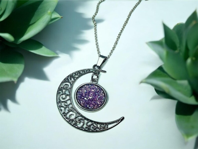 K&ouml;nnte beinhalten: Eine silberne Halbmonds-Halskette mit einem Anh&auml;nger, der mit violettem Glitzer gef&uuml;llt ist. Der Anh&auml;nger ist mit einer silbernen Kette am Mond befestigt.