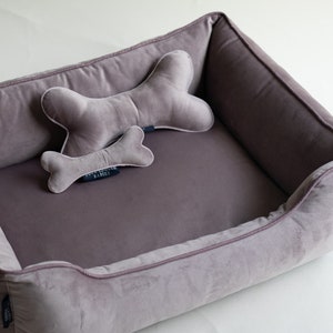 May include: A plush dog bed with a gray velvet exterior and a matching gray bone-shaped pillow. The bed has a raised rim and a soft, comfortable interior.