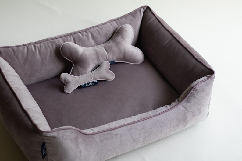 May include: A plush dog bed with a gray velvet exterior and a matching gray bone-shaped pillow. The bed has a raised rim and a soft, comfortable interior.