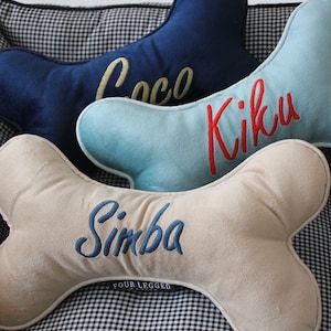 May include: Three bone-shaped dog beds with personalized names. The top bed is navy blue with the name "Coco" embroidered in gold. The middle bed is light blue with the name "Kiku" embroidered in red. The bottom bed is beige with the name "Simba" embroidered in blue. The beds are on a black and white checkered surface.