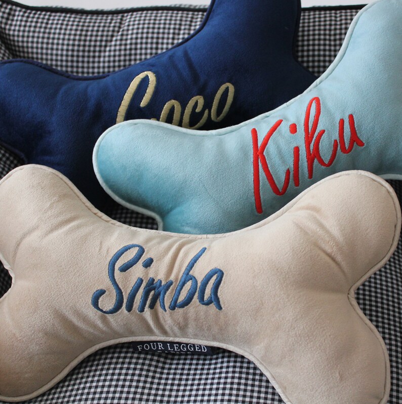 May include: Three bone-shaped dog beds with personalized names. The top bed is navy blue with the name "Coco" embroidered in gold. The middle bed is light blue with the name "Kiku" embroidered in red. The bottom bed is beige with the name "Simba" embroidered in blue. The beds are on a black and white checkered surface.