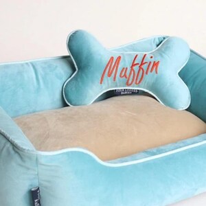 May include: A light blue dog bed with a beige cushion and a bone-shaped pillow with the name "Muffin" embroidered in orange. The bed has a white trim around the edges.