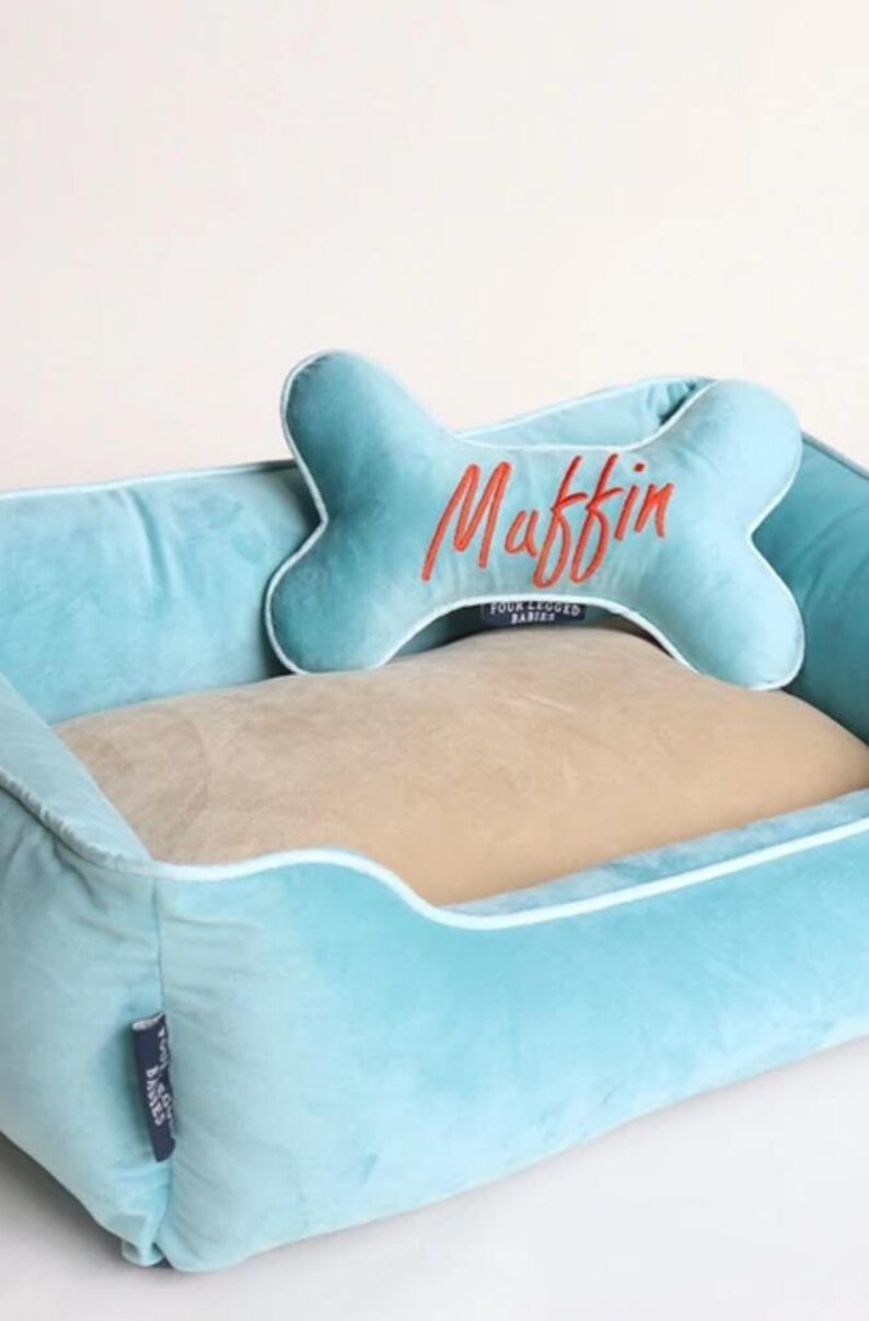 May include: A light blue dog bed with a beige cushion and a bone-shaped pillow with the name "Muffin" embroidered in orange. The bed has a white trim around the edges.