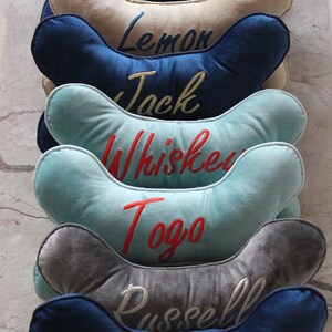 May include: A stack of six blue, gray, and light blue dog bone-shaped pillows with embroidered names: Lemon, Jack, Whiskey, Togo, and Russell.