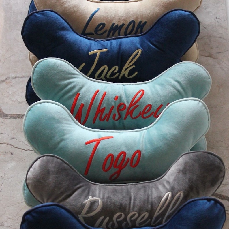 May include: A stack of six blue, gray, and light blue dog bone-shaped pillows with embroidered names: Lemon, Jack, Whiskey, Togo, and Russell.