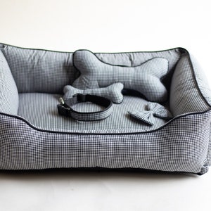 May include: A black and white checkered dog bed with a black border. The bed includes a matching bone-shaped pillow, a black collar, and a black bow tie.