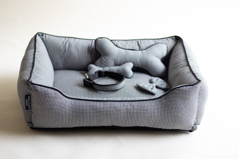 May include: A black and white checkered dog bed with a black border. The bed includes a matching bone-shaped pillow, a black collar, and a black bow tie.