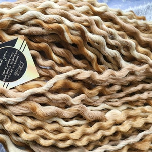 May include: A close-up of a bundle of long, curly, synthetic hair extensions in shades of blonde and brown. The hair is arranged in a loose, flowing pile. A small thank you tag is visible in the lower left corner of the image. The tag reads "Thank you for supporting our small business! You're awesome! Blessed Knitted"
