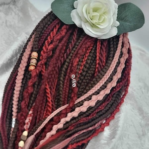 May include: A set of synthetic hair extensions in shades of red, brown, and pink. The extensions are braided and feature beads and a white flower accent.