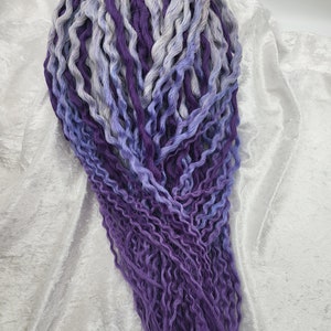 May include: A bundle of long, wavy synthetic hair extensions in shades of purple and gray. The hair is arranged in a loose, flowing style.
