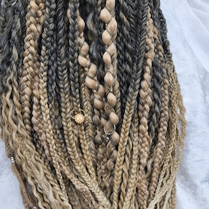 May include: Close-up of braided hair extensions in shades of brown, blonde, and gray. The braids are adorned with small gold and silver charms. The hair is styled in various textures, including twists and traditional braids.