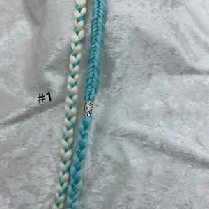 May include: Two braided hair extensions. One is light blue, the other is white with light blue accents. A silver-colored decorative bead is on the light blue braid. The number "#1" is printed in the upper left corner.